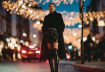Model on a city street at night under festive lights, wearing a long black coat, leather mini skirt, and black knee-high boots