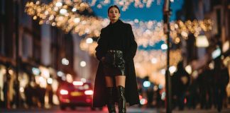 Model on a city street at night under festive lights, wearing a long black coat, leather mini skirt, and black knee-high boots