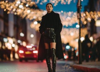 Model on a city street at night under festive lights, wearing a long black coat, leather mini skirt, and black knee-high boots