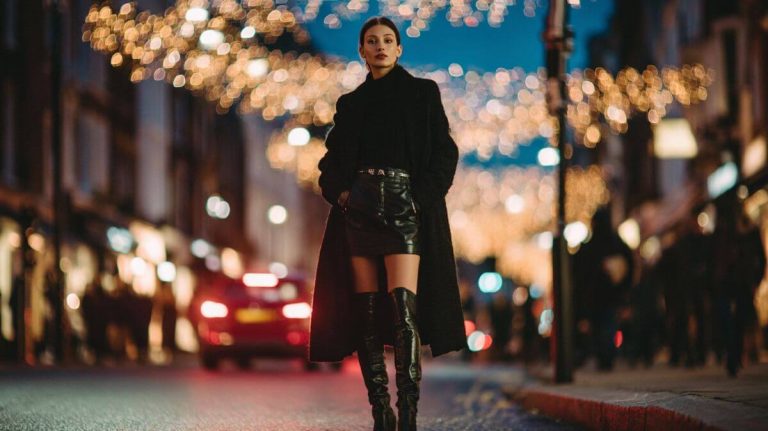 Model on a city street at night under festive lights, wearing a long black coat, leather mini skirt, and black knee-high boots